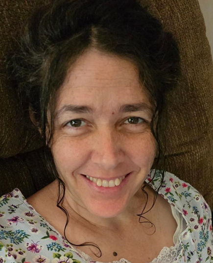 Picture of a middle-aged woman, smiling with dark brown hair spiraling down around face. She is wearing a floral top.
