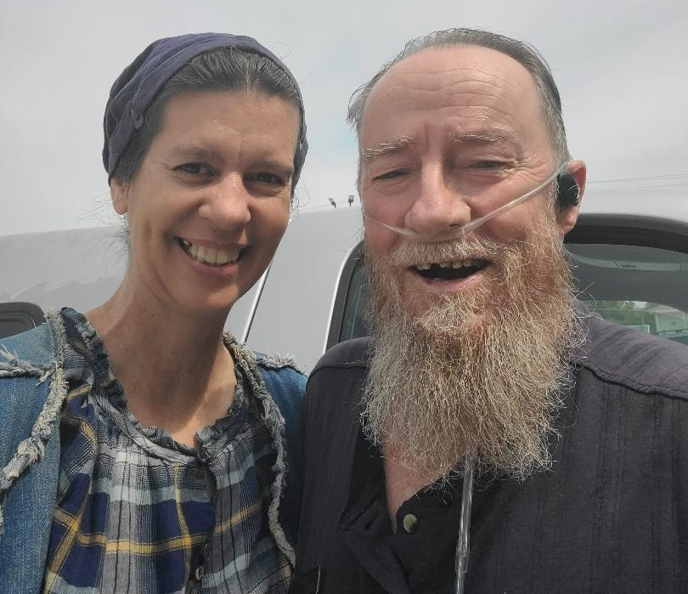 Ricky (left) wears a beard and has a portable oxygen, characterized by the hose wrapped around his face. He also has an earbud in his left ear. He is wearing a navy blue button down short-sleeved shirt. He stands beside his wife, Saige (right) who is wearing complimentary colors and a navy headcovering. She wears a denim vest over her navy plaid blouse. they are standing in front of their silver FORD truck in the thrift store parking lot.